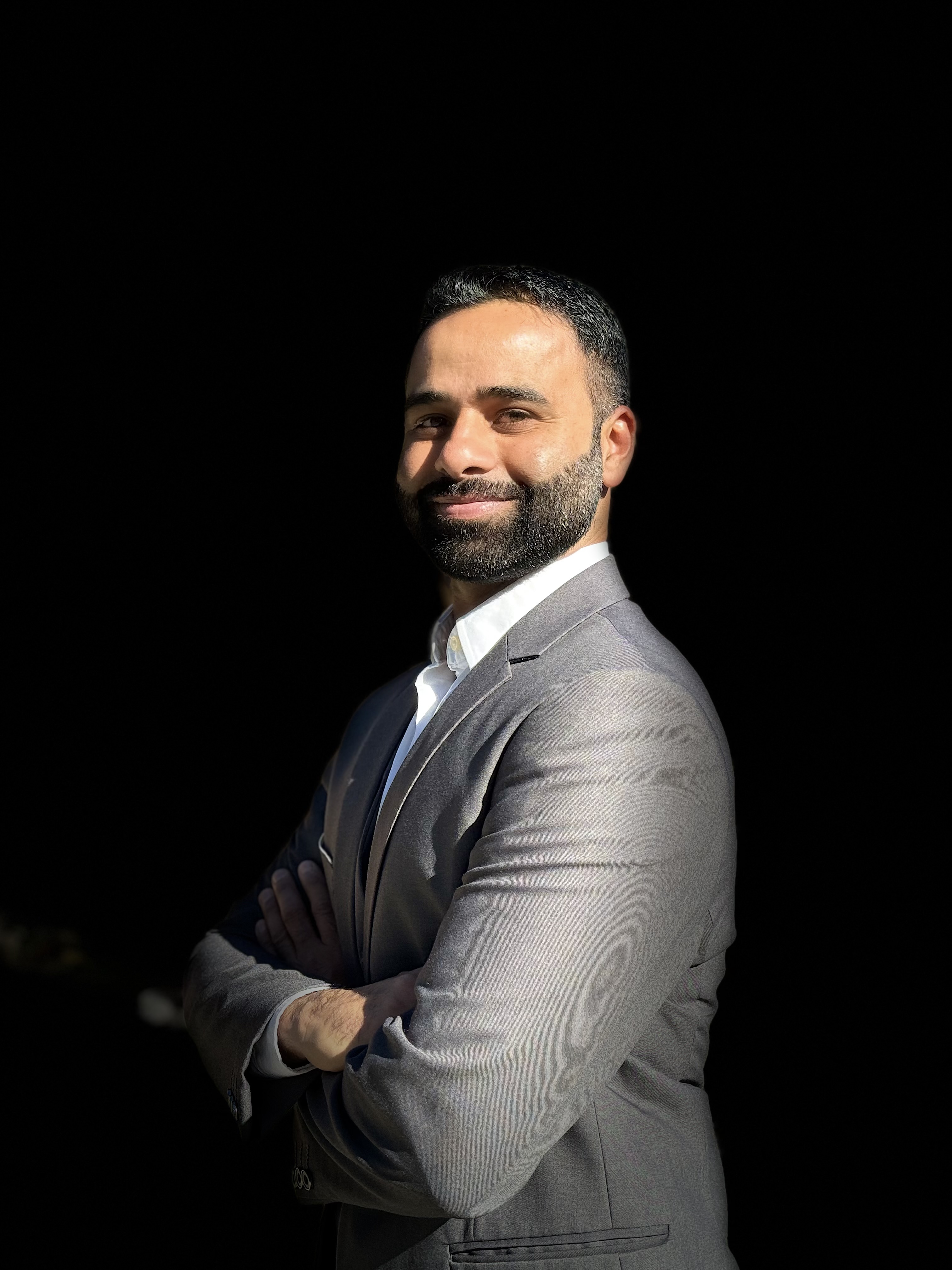 A professional portrait of a male realtor in a gray suit, standing with arms crossed, smiling confidently against a black background